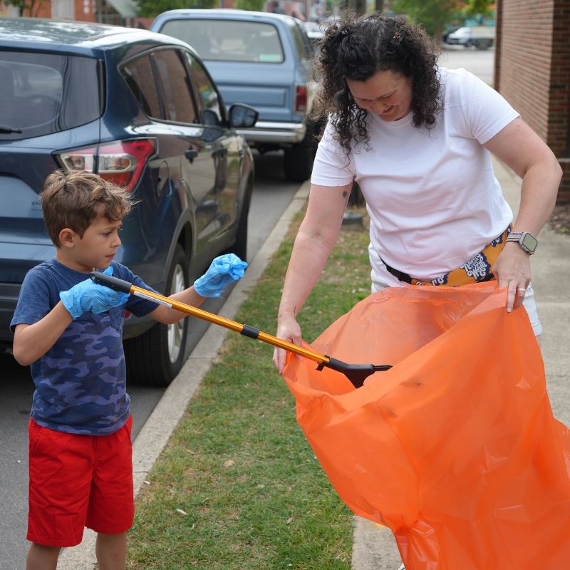 Boy uses tongs to place trash in orange trash bag that his mother is holding open